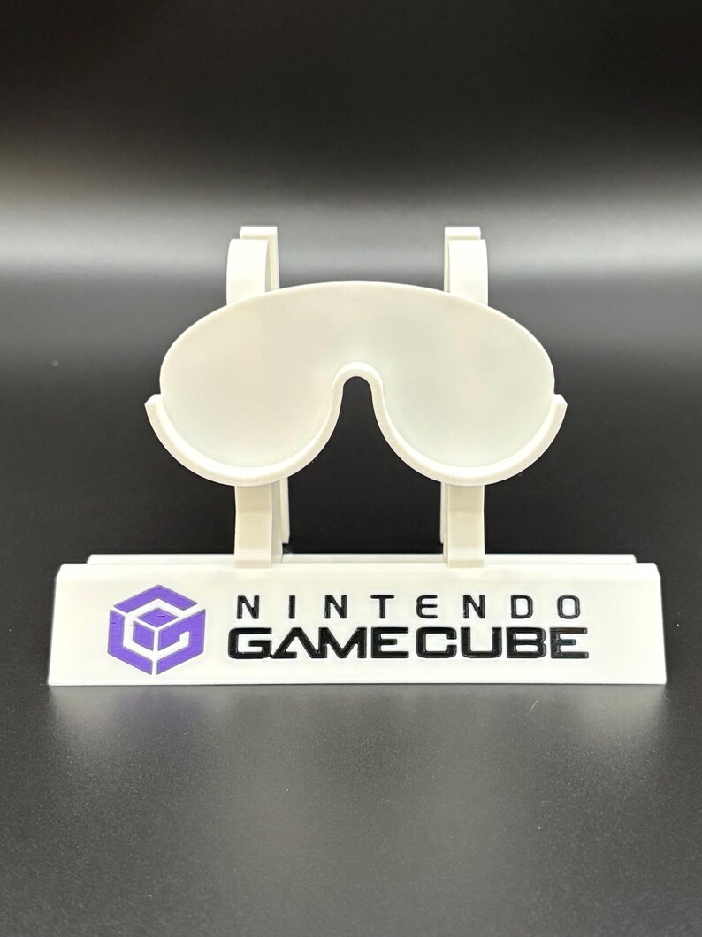 May include: A white plastic Nintendo GameCube controller stand. The stand features a stylized design with a purple GameCube logo and the words "NINTENDO GAMECUBE" in black. The stand is designed to hold a controller.