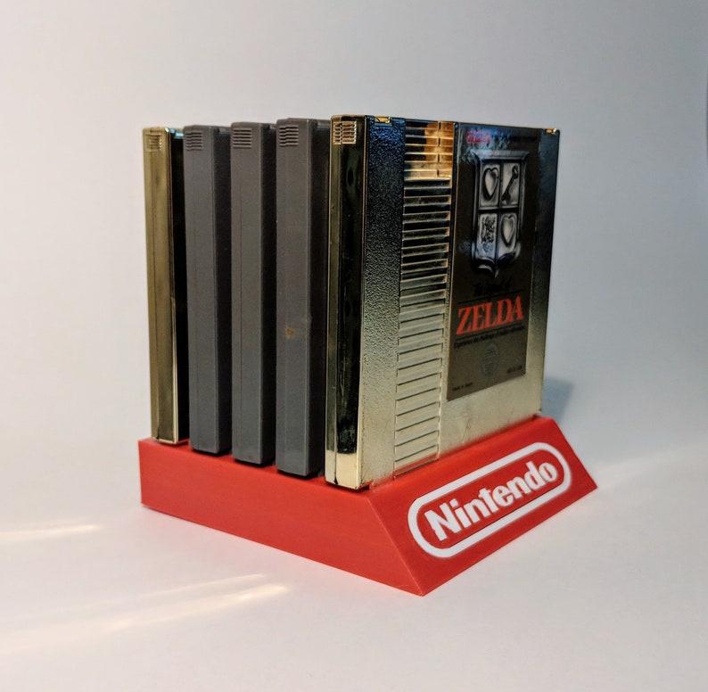 Op de afbeelding: Een rode plastic standaard met het woord "Nintendo" in wit op de voorkant. De standaard houdt vijf videospelcartridges vast, waaronder &eacute;&eacute;n gouden cartridge met het woord "Zelda" erop.