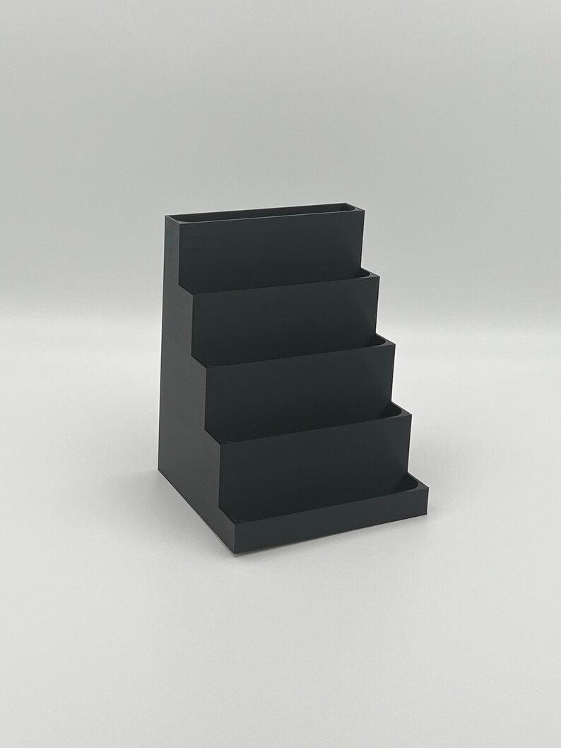 May include: A black plastic desktop organizer with four tiers. The organizer is designed to hold pens, pencils, and other office supplies.