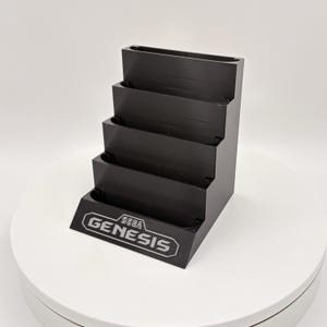 K&ouml;nnte beinhalten: Schwarzer, mehrstufiger Displayst&auml;nder f&uuml;r Sega Genesis-Spielmodule. Der St&auml;nder hat vier Ebenen und zeigt das Sega Genesis-Logo in Wei&szlig; auf der Vorderseite. Der St&auml;nder besteht aus einem mattschwarzen Material.