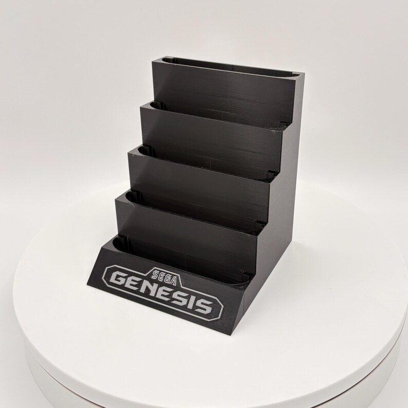 K&ouml;nnte beinhalten: Schwarzer, mehrstufiger Displayst&auml;nder f&uuml;r Sega Genesis-Spielmodule. Der St&auml;nder hat vier Ebenen und zeigt das Sega Genesis-Logo in Wei&szlig; auf der Vorderseite. Der St&auml;nder besteht aus einem mattschwarzen Material.