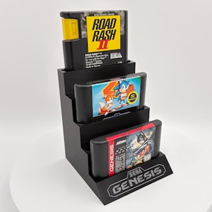K&ouml;nnte beinhalten: Ein schwarzer Displayst&auml;nder mit drei Retro-Videospielkassetten. Die oberste Kassette tr&auml;gt die Aufschrift "ROAD RASH II", die mittlere zeigt Sonic the Hedgehog und die untere "Batman Forever". Der St&auml;nder hat ein "SEGA GENESIS"-Logo.