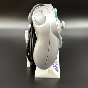 May include: Silver GameCube controller with a black cable, resting on a white stand. The controller has a distinctive button layout, including a green A button and a gray joystick. The stand has a purple logo.