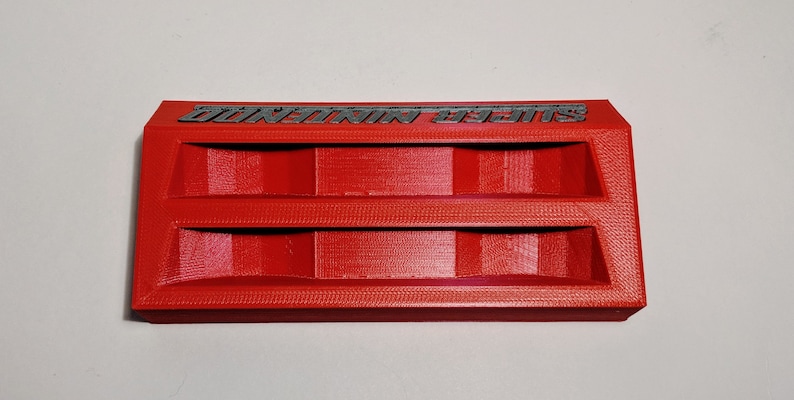 May include: A red 3D printed storage organizer with two compartments. The organizer has the text "SUPER MARIO" printed on the top.