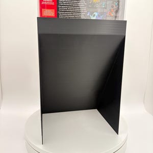 May include: A black display stand, likely for a video game case, is shown on a white rotating platform. The stand is made of a matte black material and has a unique angled design. The back of a video game case is visible.