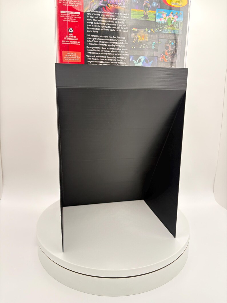 May include: A black display stand, likely for a video game case, is shown on a white rotating platform. The stand is made of a matte black material and has a unique angled design. The back of a video game case is visible.