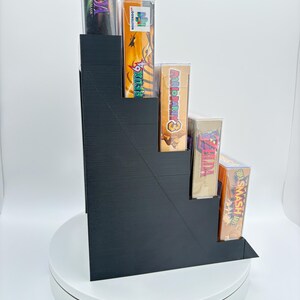 May include: A black, stepped display stand holding several video game cases. The cases feature colorful game art and titles, including "Zelda: Majora's Mask," "Star Fox 64," and "Super Smash Bros." The stand is on a white rotating platform.