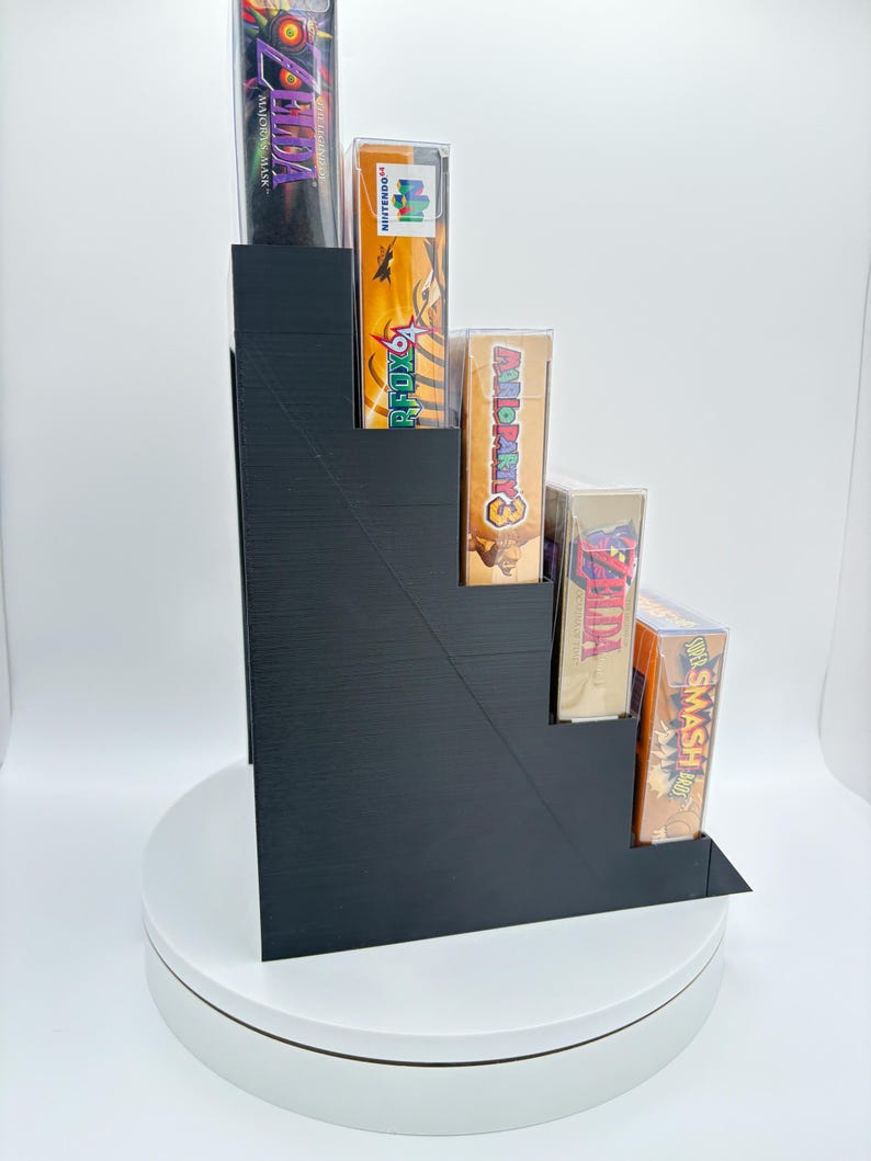 May include: A black, stepped display stand holding several video game cases. The cases feature colorful game art and titles, including "Zelda: Majora's Mask," "Star Fox 64," and "Super Smash Bros." The stand is on a white rotating platform.