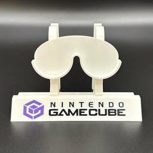 May include: A white plastic Nintendo GameCube controller stand. The stand features a stylized design to hold the controller, with the Nintendo GameCube logo and text in black and purple.