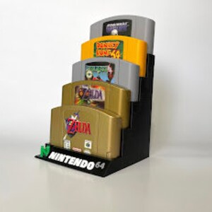 Puede incluir: Un soporte de pl&aacute;stico negro para guardar cartuchos de juegos de Nintendo 64. El soporte tiene capacidad para cuatro cartuchos, siendo el cartucho superior gris con una etiqueta blanca, el segundo cartucho verde con una etiqueta amarilla, el tercer cartucho verde con una etiqueta blanca y el cartucho inferior dorado con una etiqueta blanca. El soporte tiene la inscripci&oacute;n "NINTENDO 64" impresa en la parte delantera.