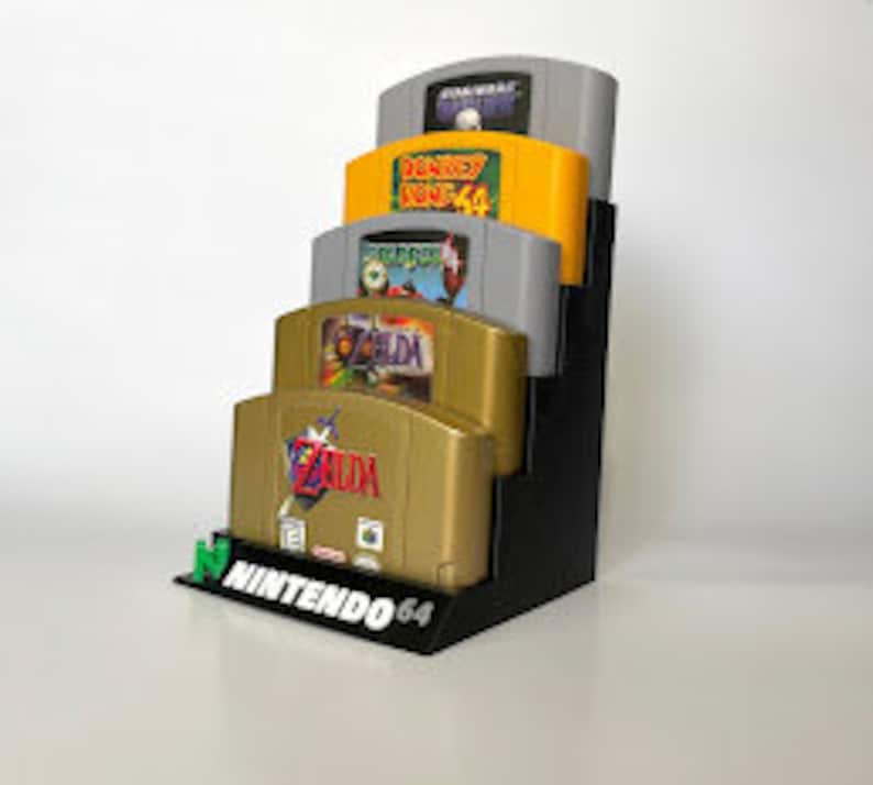 Puede incluir: Un soporte de pl&aacute;stico negro para guardar cartuchos de juegos de Nintendo 64. El soporte tiene capacidad para cuatro cartuchos, siendo el cartucho superior gris con una etiqueta blanca, el segundo cartucho verde con una etiqueta amarilla, el tercer cartucho verde con una etiqueta blanca y el cartucho inferior dorado con una etiqueta blanca. El soporte tiene la inscripci&oacute;n "NINTENDO 64" impresa en la parte delantera.