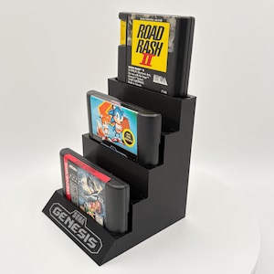 K&ouml;nnte beinhalten: Ein schwarzer, stufenf&ouml;rmiger Pr&auml;sentationsst&auml;nder mit drei Sega Genesis Videospielkassetten. Die oberste Kassette ist "Road Rash II", die mittlere ist "Sonic the Hedgehog" und die untere ist "Ghouls 'N Ghosts". Der St&auml;nder hat ein "Genesis"-Logo.