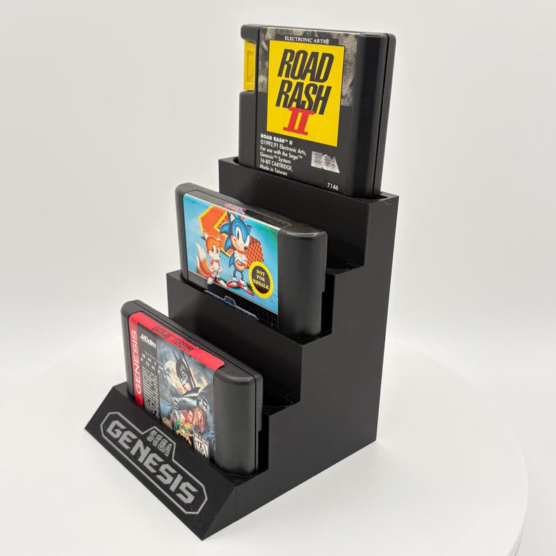 K&ouml;nnte beinhalten: Ein schwarzer, stufenf&ouml;rmiger Pr&auml;sentationsst&auml;nder mit drei Sega Genesis Videospielkassetten. Die oberste Kassette ist "Road Rash II", die mittlere ist "Sonic the Hedgehog" und die untere ist "Ghouls 'N Ghosts". Der St&auml;nder hat ein "Genesis"-Logo.