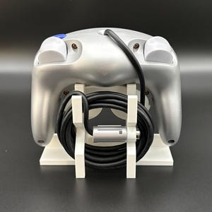 May include: Silver-colored video game controller with a black cable, resting on a white stand. The controller has two white buttons and one blue button. The stand is white and has a minimalist design.