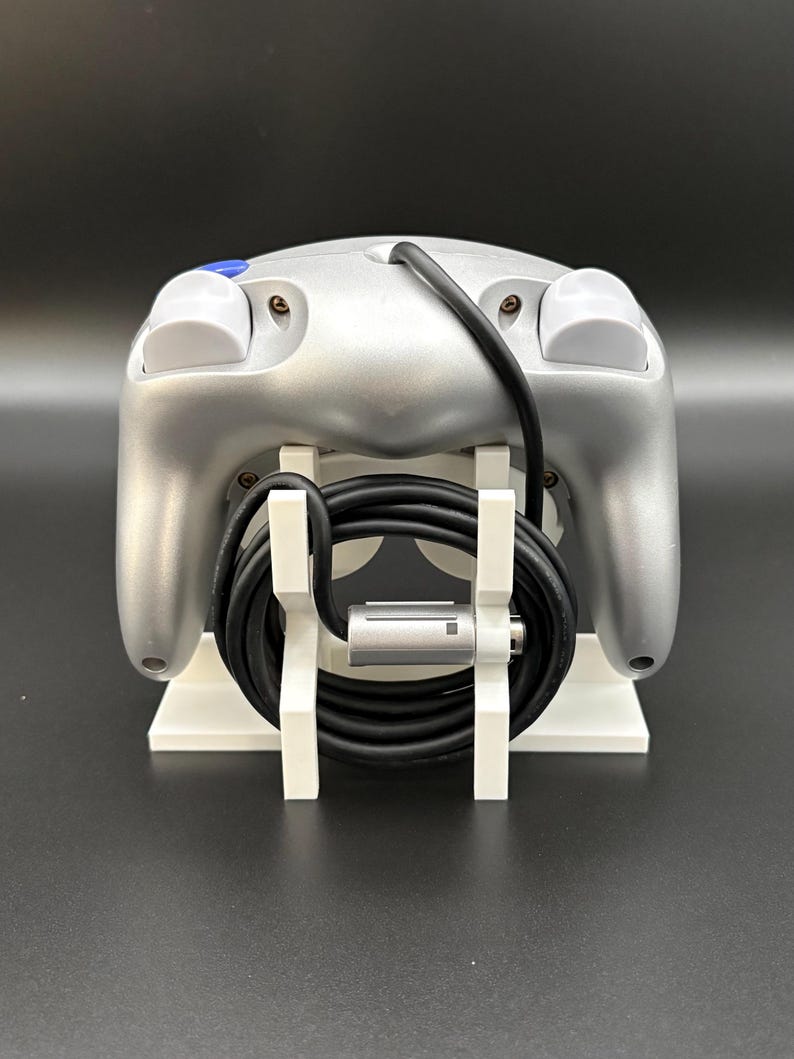 May include: Silver-colored video game controller with a black cable, resting on a white stand. The controller has two white buttons and one blue button. The stand is white and has a minimalist design.
