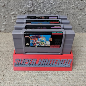 May include: A red plastic stand with the text "SUPER NINTENDO ENTERTAINMENT SYSTEM" printed on the front. Four gray Super Nintendo game cartridges are stacked in the stand. The cartridges are labeled "Super Mario World", "Yoshi's Island", "Mario Paint", and "Super Mario World 2: Yoshi's Island".