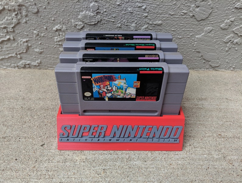 May include: A red plastic stand with the text "SUPER NINTENDO ENTERTAINMENT SYSTEM" printed on the front. Four gray Super Nintendo game cartridges are stacked in the stand. The cartridges are labeled "Super Mario World", "Yoshi's Island", "Mario Paint", and "Super Mario World 2: Yoshi's Island".