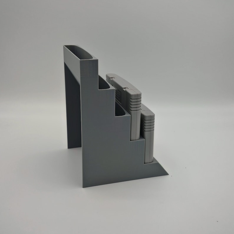 Puede incluir: Un organizador gris escalonado dise&ntilde;ado para guardar cartuchos de videojuegos. El organizador tiene una base rectangular y una secci&oacute;n vertical alta. Dos cartuchos de juego grises est&aacute;n colocados en el organizador. El dise&ntilde;o es simple y moderno.