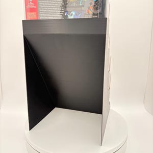 May include: Black display stand with a black front and sides, showcasing a video game box. The stand is on a white rotating platform. The video game box has colorful graphics and text.