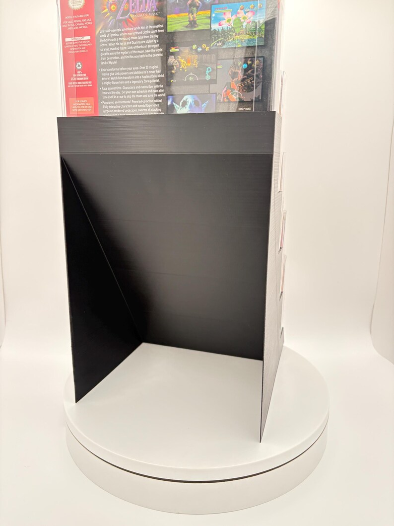 May include: Black display stand with a black front and sides, showcasing a video game box. The stand is on a white rotating platform. The video game box has colorful graphics and text.
