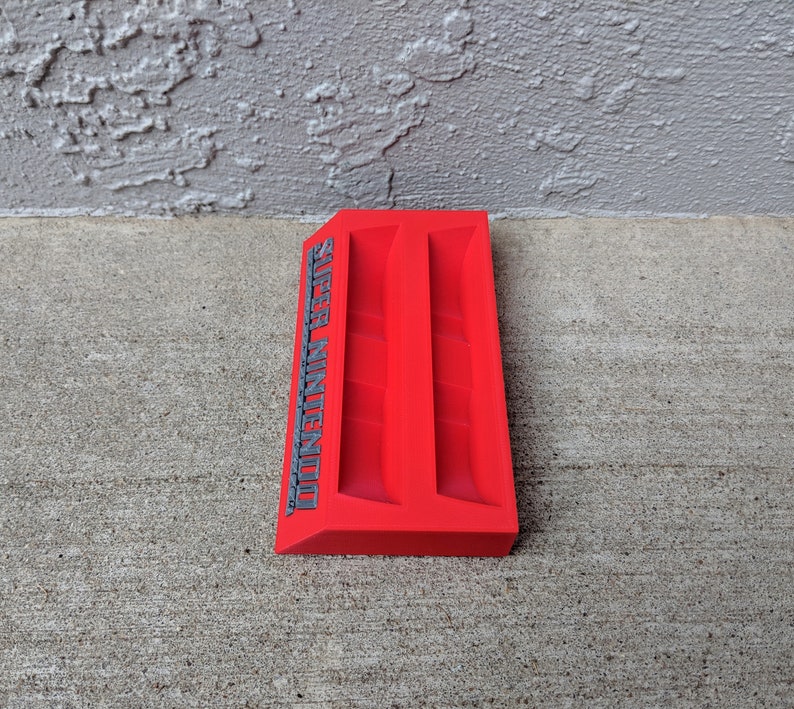 May include: Red plastic Super Nintendo game cartridge holder with two slots. The holder has the words "Super Nintendo" printed on the side in gray.