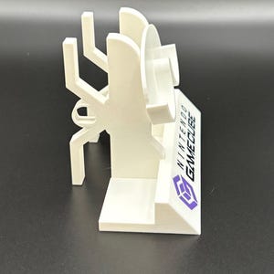May include: A white 3D-printed stand for a Nintendo GameCube console. The stand features a stylized design with the GameCube logo and text. The stand is designed to hold the console and games.