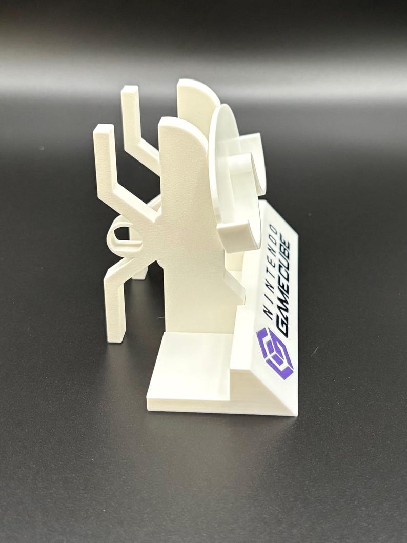 May include: A white 3D-printed stand for a Nintendo GameCube console. The stand features a stylized design with the GameCube logo and text. The stand is designed to hold the console and games.