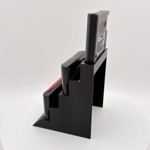 K&ouml;nnte beinhalten: Schwarzer 3D-gedruckter Spielkassettenhalter mit drei Schlitzen, der zwei schwarze Spielkassetten mit rot-wei&szlig;en Etiketten h&auml;lt. Der Halter ist so konzipiert, dass die Kassetten in einer abgestuften Anordnung angezeigt werden.