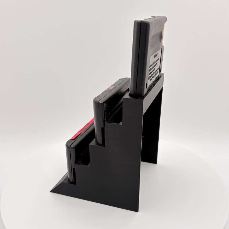 K&ouml;nnte beinhalten: Schwarzer 3D-gedruckter Spielkassettenhalter mit drei Schlitzen, der zwei schwarze Spielkassetten mit rot-wei&szlig;en Etiketten h&auml;lt. Der Halter ist so konzipiert, dass die Kassetten in einer abgestuften Anordnung angezeigt werden.