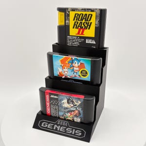 K&ouml;nnte beinhalten: Ein schwarzer St&auml;nder mit drei Sega Genesis-Spielkassetten. Die oberste Kassette ist "Road Rash II", die mittlere zeigt Sonic the Hedgehog und die untere "Batman Forever". Der St&auml;nder hat das Sega Genesis-Logo.