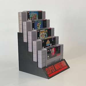 Puede incluir: Un expositor negro para cartuchos de juegos de Super Nintendo Entertainment System con cinco cartuchos de juegos. Los cartuchos est&aacute;n etiquetados como "Super Mario World", "Super Mario All-Stars", "Super Mario Kart", "Super Metroid" y "Final Fantasy VI".