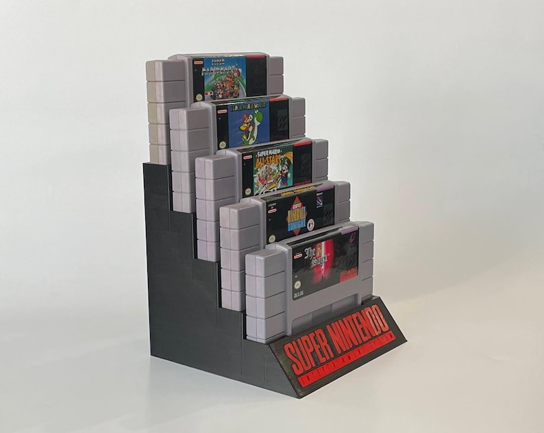 Puede incluir: Un expositor negro para cartuchos de juegos de Super Nintendo Entertainment System con cinco cartuchos de juegos. Los cartuchos est&aacute;n etiquetados como "Super Mario World", "Super Mario All-Stars", "Super Mario Kart", "Super Metroid" y "Final Fantasy VI".