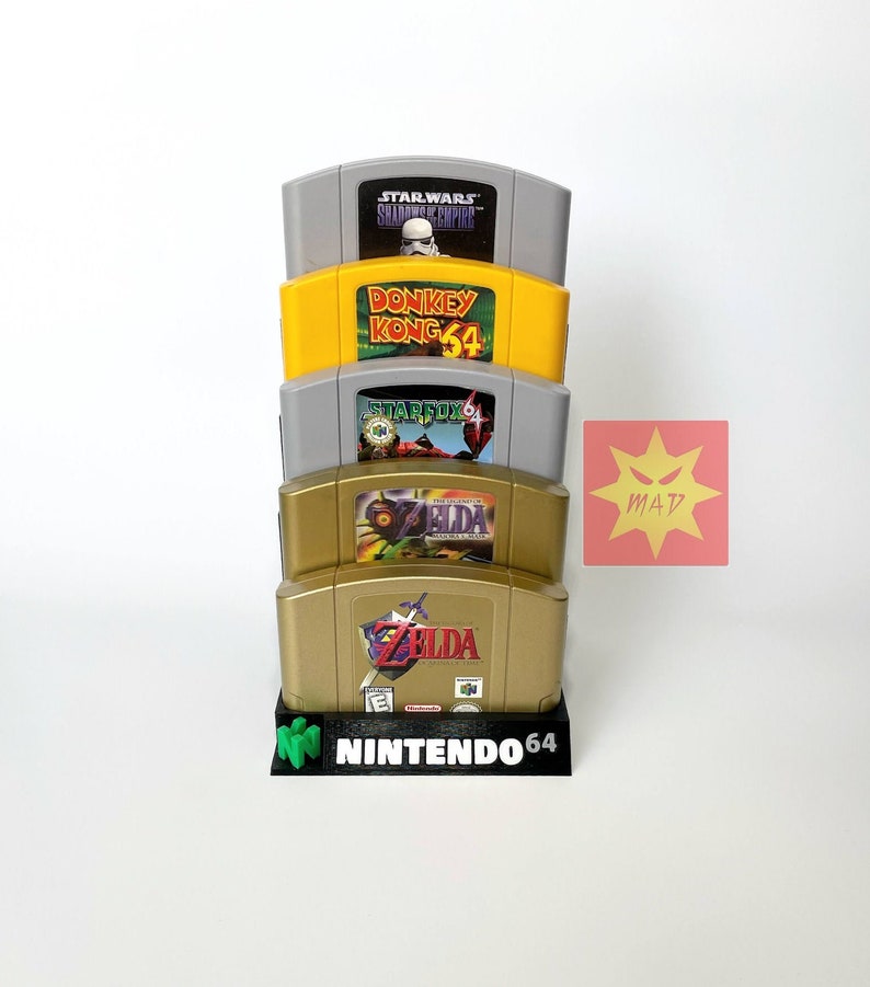 Puede incluir: Una pila de cinco cartuchos de juego de Nintendo 64 en un soporte. Los cartuchos son, de arriba a abajo: Star Wars: Shadows of the Empire, Donkey Kong 64, Star Fox 64, The Legend of Zelda: Ocarina of Time y The Legend of Zelda: Majora&rsquo;s Mask.