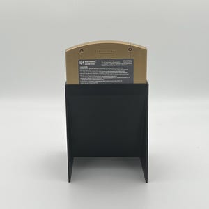 May include: A gold Nintendo 64 game cartridge for the game "GoldenEye 007" is displayed in a black plastic stand. The cartridge has the Nintendo logo and the game title printed on it.