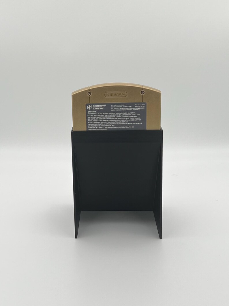 May include: A gold Nintendo 64 game cartridge for the game "GoldenEye 007" is displayed in a black plastic stand. The cartridge has the Nintendo logo and the game title printed on it.
