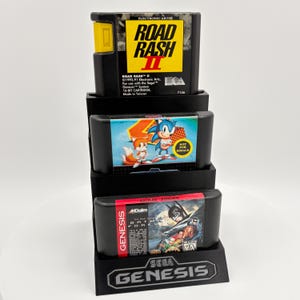 K&ouml;nnte beinhalten: Ein schwarzer Displayst&auml;nder mit drei Sega Genesis-Spielkassetten. Die oberste Kassette ist "Road Rash II", die mittlere zeigt Sonic the Hedgehog und die untere "Batman Forever". Der St&auml;nder hat das Sega Genesis-Logo.