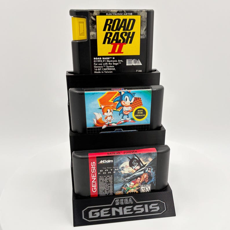 K&ouml;nnte beinhalten: Ein schwarzer Displayst&auml;nder mit drei Sega Genesis-Spielkassetten. Die oberste Kassette ist "Road Rash II", die mittlere zeigt Sonic the Hedgehog und die untere "Batman Forever". Der St&auml;nder hat das Sega Genesis-Logo.