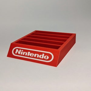 Op de afbeelding: Rode plastic Nintendo-spelcartridgehouder met zes slots. De houder heeft een wit Nintendo-logo aan de voorkant.