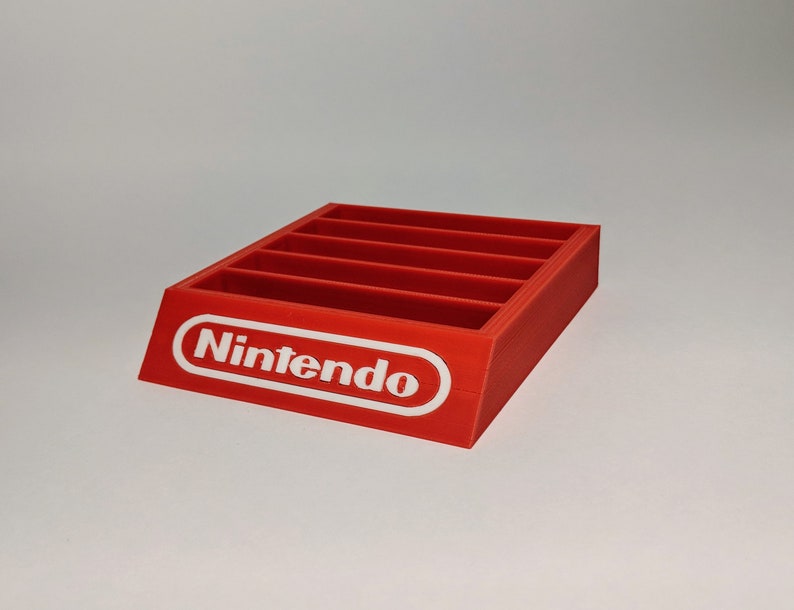Op de afbeelding: Rode plastic Nintendo-spelcartridgehouder met zes slots. De houder heeft een wit Nintendo-logo aan de voorkant.