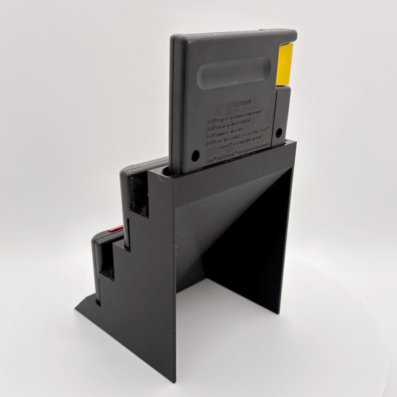 K&ouml;nnte beinhalten: Ein schwarzer, 3D-gedruckter Spielkassettenhalter mit eingelegter Kassette. Der Halter hat ein Stufendesign auf einer Seite und eine dreieckige St&uuml;tze. Die Kassette ist schwarz mit einem gelben Tab.