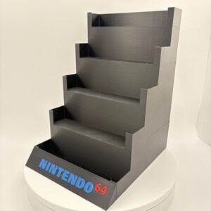 May include: Black tiered display stand with the text "NINTENDO 64" in blue and red. The stand has five levels, each with a small lip to hold items. The stand is designed to display video game cartridges or other collectibles.