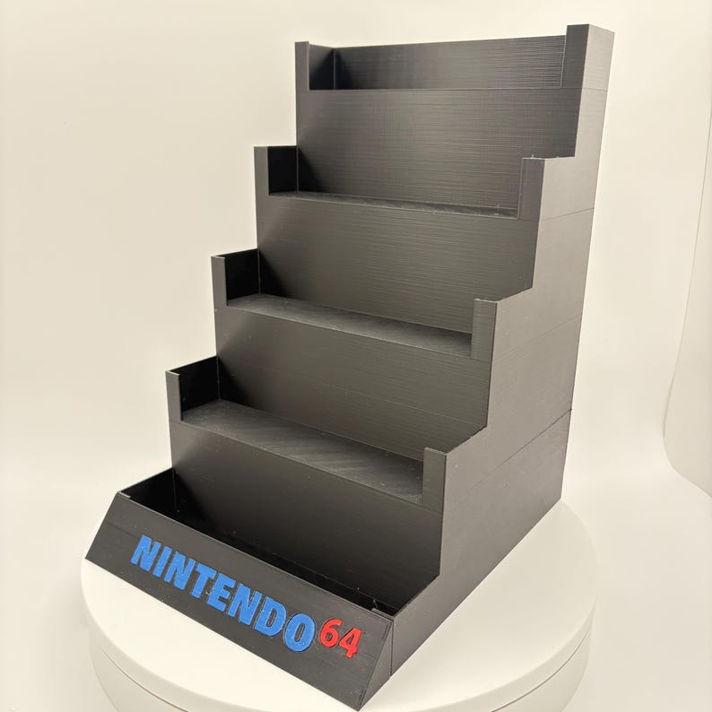 May include: Black tiered display stand with the text "NINTENDO 64" in blue and red. The stand has five levels, each with a small lip to hold items. The stand is designed to display video game cartridges or other collectibles.