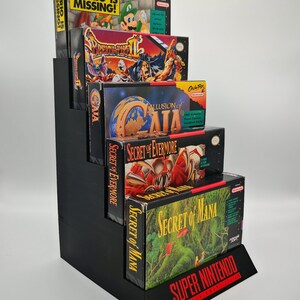 May include: A black, stepped display stand holding five vintage Super Nintendo video game boxes. The boxes feature colorful game art and titles like "Mario is Missing!" and "Secret of Mana." The stand has a "Super Nintendo" logo.