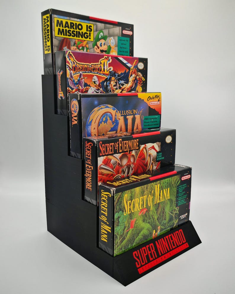 May include: A black, stepped display stand holding five vintage Super Nintendo video game boxes. The boxes feature colorful game art and titles like "Mario is Missing!" and "Secret of Mana." The stand has a "Super Nintendo" logo.