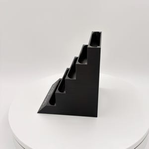 K&ouml;nnte beinhalten: Ein schwarzer, stufenf&ouml;rmiger Pr&auml;sentationsst&auml;nder mit vier Ebenen. Jede Ebene hat einen gebogenen, vertieften Bereich. Der St&auml;nder ist abgewinkelt, mit einem dekorativen Design an der Seite. Der St&auml;nder besteht aus einem matten Material.
