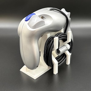 May include: Silver video game controller with a blue button and a black cable, resting on a white stand. The controller has a sleek design and is likely for a gaming console. The stand neatly organizes the controller and its cable.