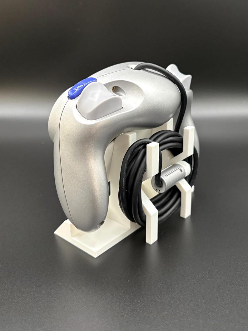 May include: Silver video game controller with a blue button and a black cable, resting on a white stand. The controller has a sleek design and is likely for a gaming console. The stand neatly organizes the controller and its cable.