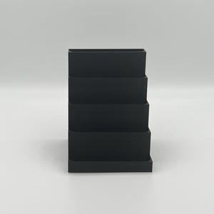 May include: A black plastic tiered organizer with four compartments. The organizer is designed to hold small items like pens, pencils, or other office supplies.