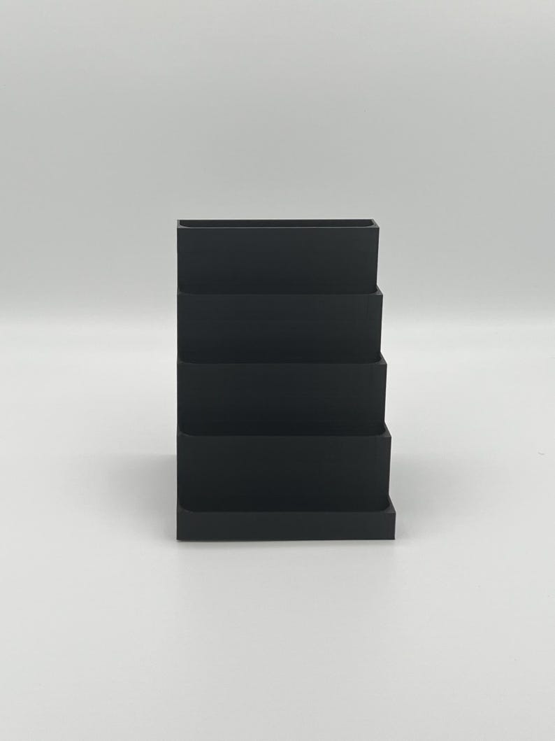 May include: A black plastic tiered organizer with four compartments. The organizer is designed to hold small items like pens, pencils, or other office supplies.