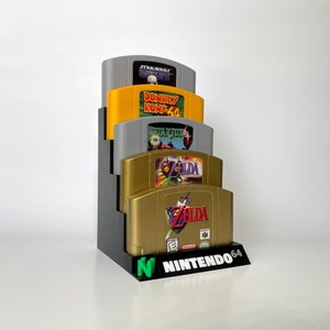 Puede incluir: Un soporte negro para cartuchos de juegos de Nintendo 64 con cuatro cartuchos de juegos. Los cartuchos son, de arriba a abajo: Star Wars Shadows of the Empire, Donkey Kong 64, Starfox 64 y The Legend of Zelda: Ocarina of Time.